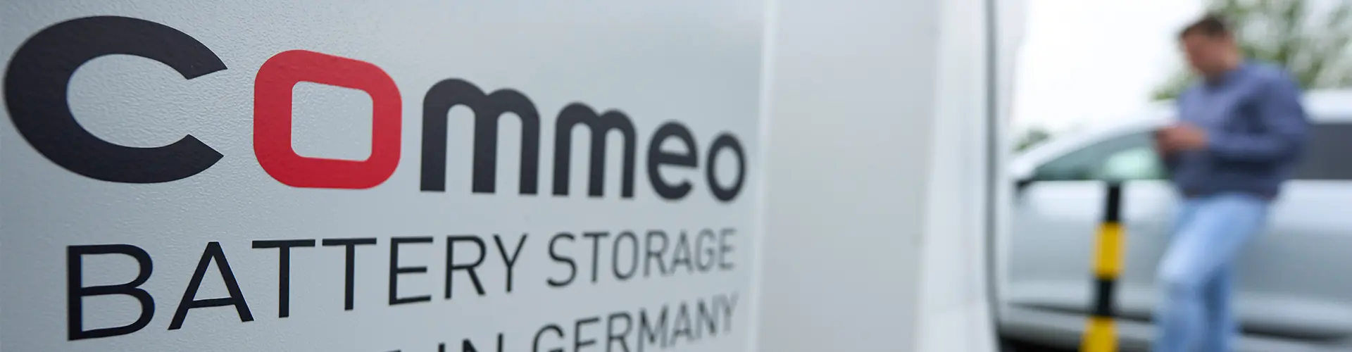 Das Bild präsentiert einen Commeo Batteriespeicher mit der Aufschrift "Commeo Battery Storage - Hergestellt in Deutschland". Im Hintergrund ist ein Herr zu erkennen, der sein Elektrofahrzeug lädt.