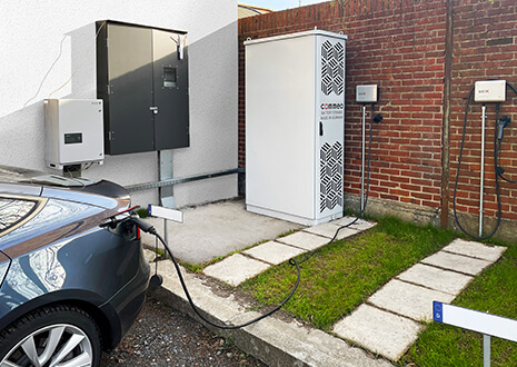 The picture presents a Commeo battery storage in the outdoor area of the company bill-x GmbH. Clearly visible is an e-car that is being charged by this storage.