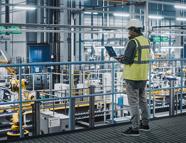 Das Bild vermittelt den Eindruck eines engagierten Fabriktechnikers, der im Stehen mit seinem Laptop arbeitet und dabei den Automatisierungsprozess aufmerksam überwacht.